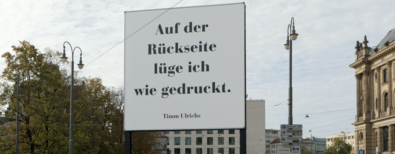 Diagonal view of the billboard. To be read in large black letters on a white background: "Auf der Rückseite lüge ich wie gedruckt. Timm Ulrichs" ("I lie as printed on the reverse. Timm Ulrichs"